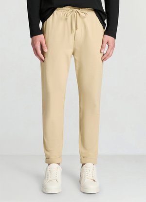 Beige Jogginghosen mit elastischem Bund und Kordelzug. Weicher Stoff mit glatter Textur, verjüngt sich zu den Bündchen hin. Kombiniert mit weißen Sneakers.