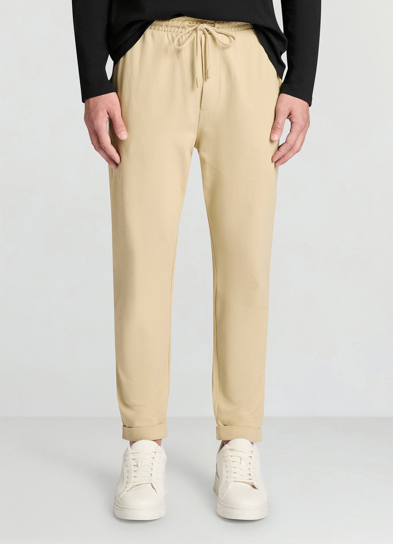 Beige Jogginghosen mit elastischem Bund und Kordelzug. Weicher Stoff mit glatter Textur, verjüngt sich zu den Bündchen hin. Kombiniert mit weißen Sneakers.