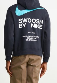 Marinblå hoodie med stor vit text "SWOOSH BY NIKE" och blå swooshdetalj på baksidan. Mjuk bomullstextil med ribbad nederkant och ärmslut.