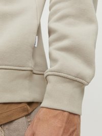 Beige Sweatshirtstoff mit gerippten Bündchen und einem kleinen Markenlabel an der Seite. Die Textur wirkt weich und glatt.