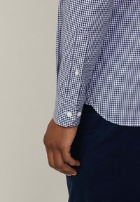 Navy and white checkered shirt with buttoned cuffs, featuring a curved hem and a soft cotton texture. Button accents are white and rounded.