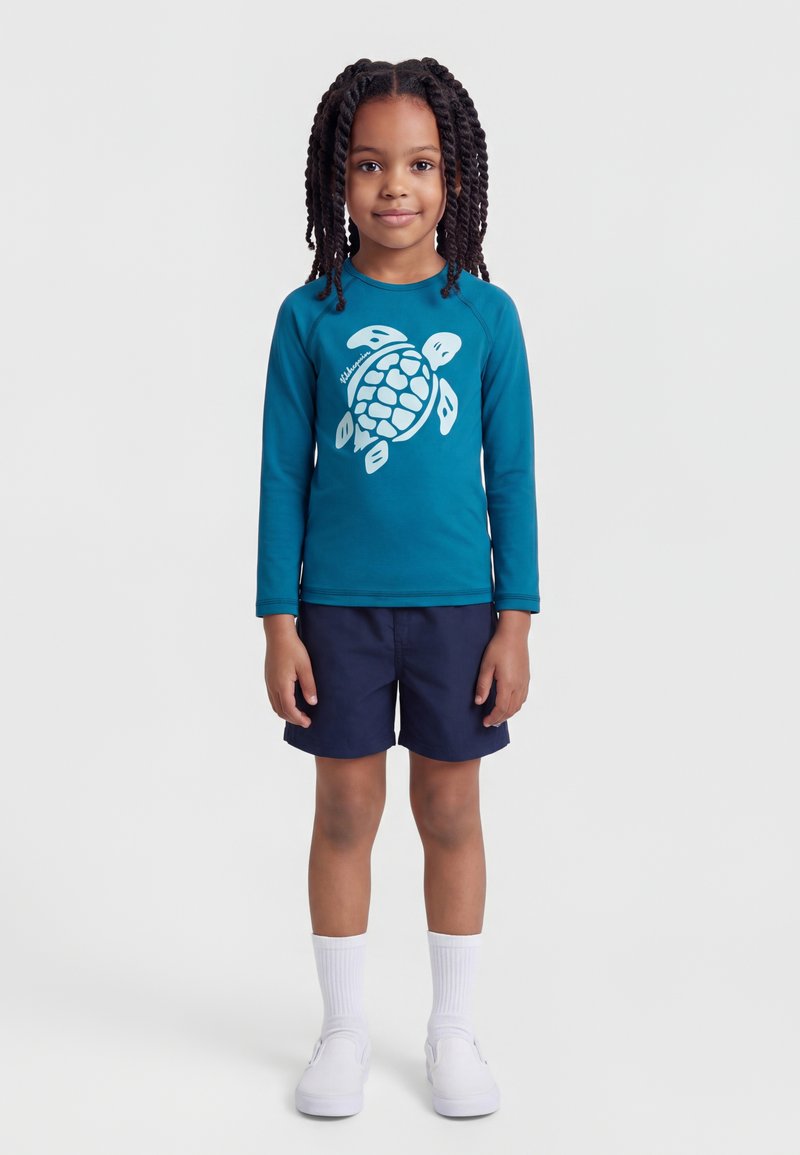 Παιδί με πλεξούδες, φοράει μπλουζάκι μακρυμάνικο σε τιρκουάζ χρώμα με σχέδιο θαλάσσιας χελώνας, σκούρο μπλε σορτς, λευκές κάλτσες και λευκά παπούτσια χωρίς κορδόνια.