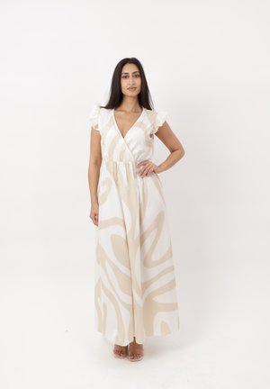 Donna in piedi con la mano sui fianchi, indossa un abito lungo senza maniche con motivo astratto beige e bianco e spalle arricciate.
