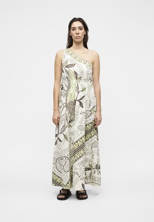 Γυναίκα που στέκεται, φορώντας ένα λευκό μακρύ φόρεμα με έναν ώμο, με πράσινα και καφέ βοτανικά σχέδια και μαύρα σανδάλια.