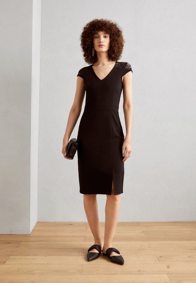 Abito nero aderente con maniche corte in pizzo a cappuccio, scollo a V, lunghezza al ginocchio con uno spacco laterale, abbinato a una borsa a tracolla nera e scarpe basse.