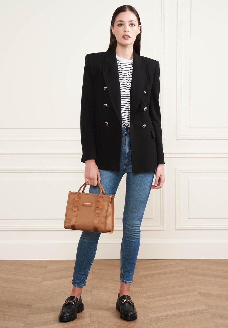 Blazer noir à double boutonnage, t-shirt rayé, jeans skinny bleus, sac fourre-tout marron avec poignées, et mocassins noirs. Fond de sol en bois plat.