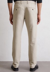 Beige cotton trousers with a straight cut, back pockets with button closures, worn with black footwear on a neutral background.