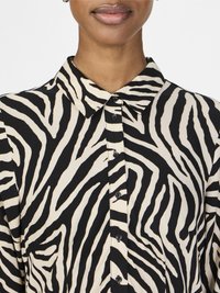 Camisa com padrão de zebra em preto e bege, com pala de botões, colarinho e uma textura de tecido suave. Apresenta riscas em contraste e um design ajustado.