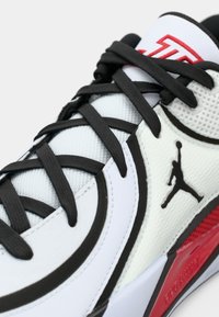 Κοντινό πλάνο λευκού και μαύρου αθλητικού παπουτσιού Air Jordan με μαύρες κορδόνες, κόκκινη λεπτομέρεια στη σόλα και λογότυπο Jumpman στο υφή πλευρικής πάνελ.