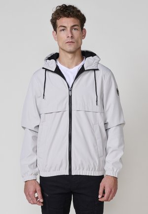 Chaqueta impermeable de color gris claro con capucha, cierre frontal con cremallera, puños elásticos y dos bolsillos laterales. Cuenta con un forro negro y detalles de acento.