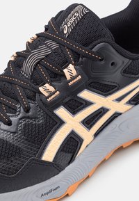 Gros plan sur une chaussure de course Asics noire avec des accents pêche, semelle texturée étiquetée AmpliFoam, et un laçage détaillé sur la tige en mesh.