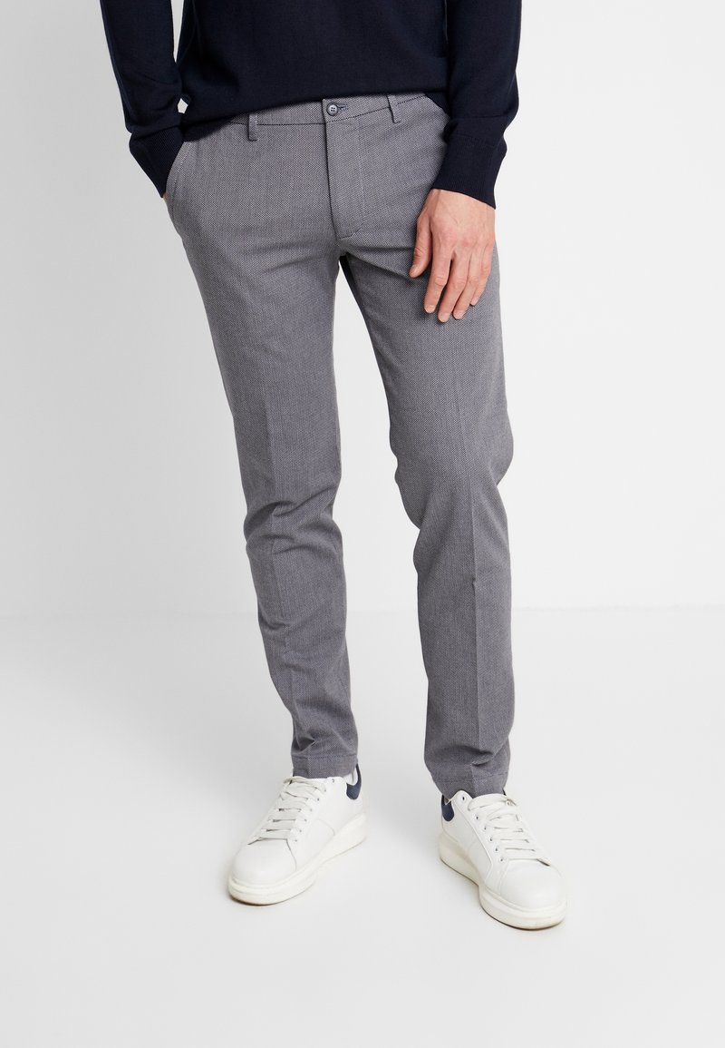 Homme portant un pantalon gris slim et des baskets blanches, debout avec une main dans la poche devant un fond uni.