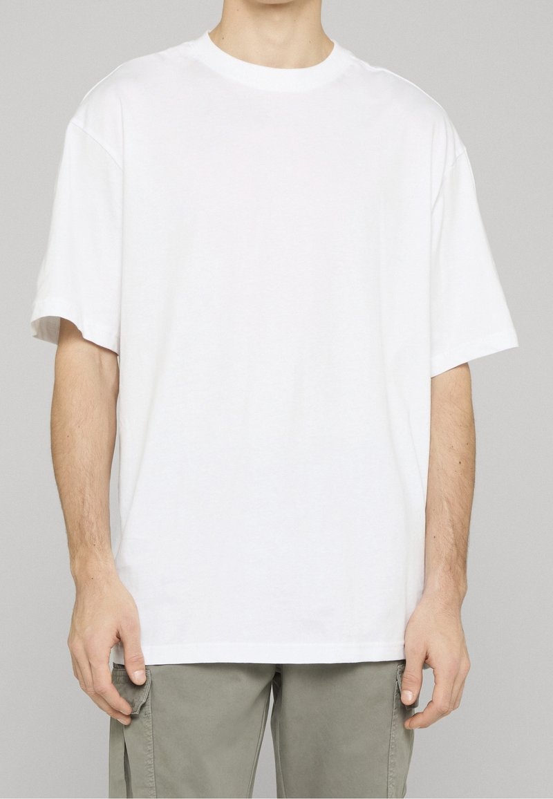 Personne portant un t-shirt blanc uni à manches courtes et oversized avec un pantalon cargo gris clair sur un fond neutre.