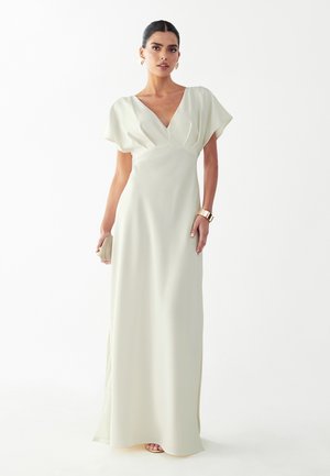 Frau trägt ein bodenlanges, weißes Kleid mit V-Ausschnitt und kurzen Ärmeln, hält eine kleine beige Clutch und trägt goldenen Schmuck.