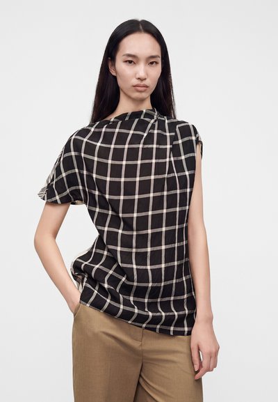 Femme aux longs cheveux noirs portant un haut à carreaux noir et blanc et un pantalon beige, debout avec une main dans la poche devant un fond blanc.
