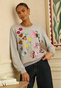 Szara bluza z haftem z cekinów w kolorze różowym, żółtym i zielonym. Mankiety i dół bluzy wykończone ściągaczami; luźny krój. Noszona z ciemnymi dżinsami.