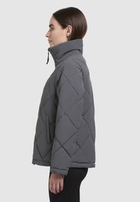 Gray quilted puffer jacket with a high collar, front pocket, and a textured finish. Features a zip closure at the neck for added warmth.