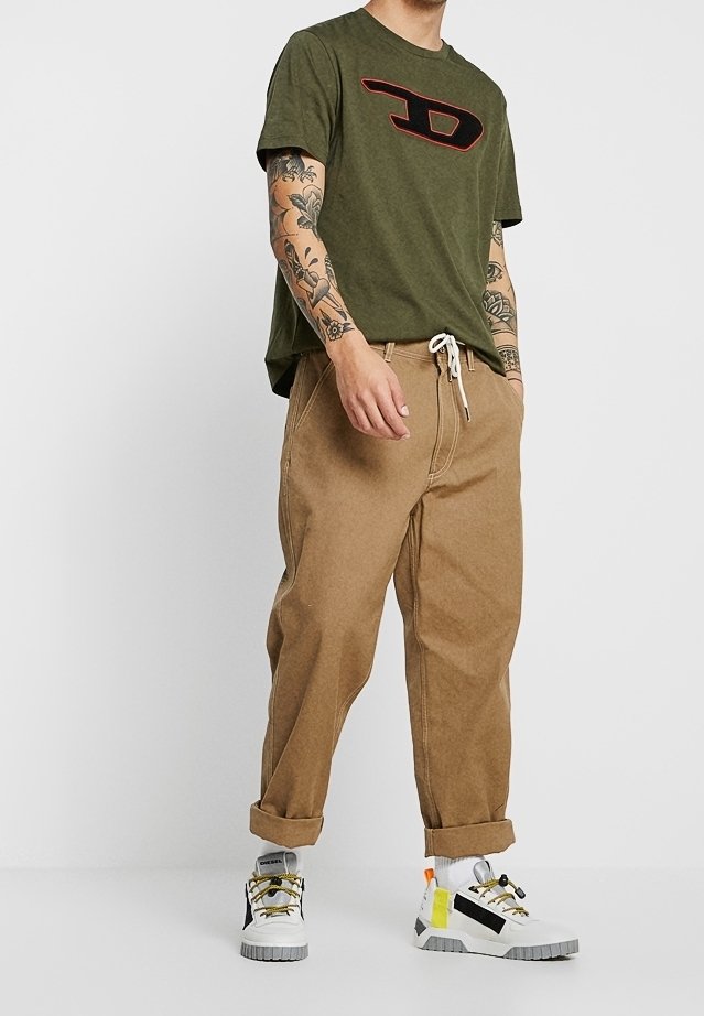 Homme portant un t-shirt vert olive avec un logo, un pantalon sable retroussé avec une taille à cordon, des baskets montantes blanches et des tatouages visibles sur les bras.