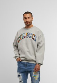 Hanorac gri cu litere multicolore "COLUCCI". Are gulerul rotund și o croială lejeră; asortat cu blugi uzati de culoare albastră.