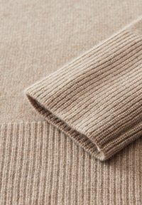 Beiger Strickstoff mit gerippter Textur, der einen umgeschlagenen Ärmelrand aufweist und eine weiche, warme Ausstrahlung hat. Nahaufnahme des Materials.