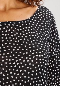 Czarna tkanina z wzorem małych białych serc noszona jako bluzka z długim rękawem i okrągłym dekoltem.