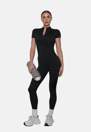 Kobieta w czarnym dopasowanym stroju sportowym trzyma teksturowany szary wałek piankowy, ma na sobie białe buty sportowe i skarpetki, stoi na białym tle.