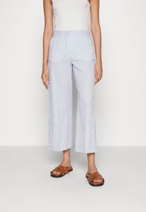 Femme portant un haut blanc sans manches, un pantalon large bleu clair à fines rayures et des sandales marron, debout devant un fond uni.