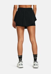 Pantaloni sportivi neri con sovrapposizione svasata, vita elastica e pantaloni interni. Indossati con sneaker bianche e calzini a righe.