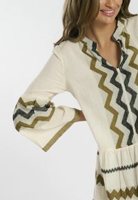 Tunique crème texturée avec un col en V et des manches à cloche. Présente des motifs en zigzag noir et vert olive le long de l'encolure et des manches.