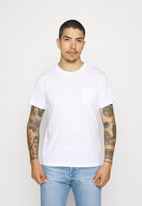 T-shirt bianco in cotone con scollatura rotonda, maniche corte e taschino sul lato sinistro del petto. Indossato con jeans in denim azzurro chiaro.