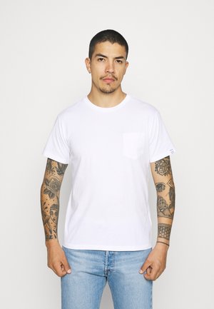 T-shirt bianco in cotone con scollatura rotonda, maniche corte e taschino sul lato sinistro del petto. Indossato con jeans in denim azzurro chiaro.
