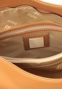 Sac en cuir fauve avec intérieur en tissu beige, comprenant une poche zippée et une étiquette métallique de marque portant l'inscription "Kazar".