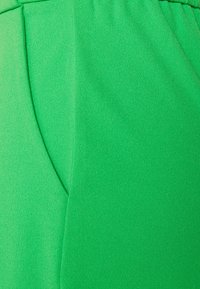 Zelená, texturovaná látka oděvu s hladkým povrchem; zahrnuje boční kapsu a detaily švů, které zdůrazňují přiléhavý design.