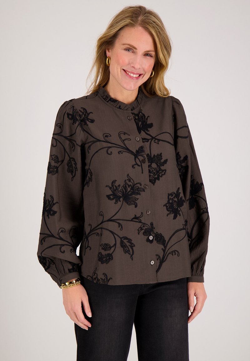 Vrouw glimlachend, gekleed in een donkerblouse met knopen, zwart bloemenborduursel en zwarte broek tegen een effen lichte achtergrond.