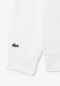 Polsino di una felpa bianca in cotone con finitura testurizzata e un piccolo logo di coccodrillo verde sulla manica sinistra.