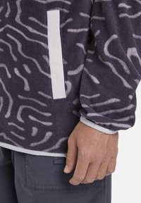Fleece pullover in dark gray with white abstract wave pattern, featuring a horizontal white accent pocket and elastic cuffs.