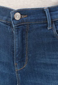 Jeans de mezclilla azul con un lavado claro, que presentan un cierre con botón plateado, acentos de costura marrón y un pasador para cinturón en la cintura.
