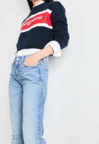 Suéter navy con franjas rojas y blancas, con el texto "Tommy Jeans". Lleva puesto sobre una camisa blanca de cuello. Combinado con jeans de color azul claro.