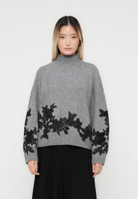 Femme portant un pull col montant gris avec des broderies florales noires sur les manches et l'ourlet, associé à une jupe plissée noire, se tenant de face.