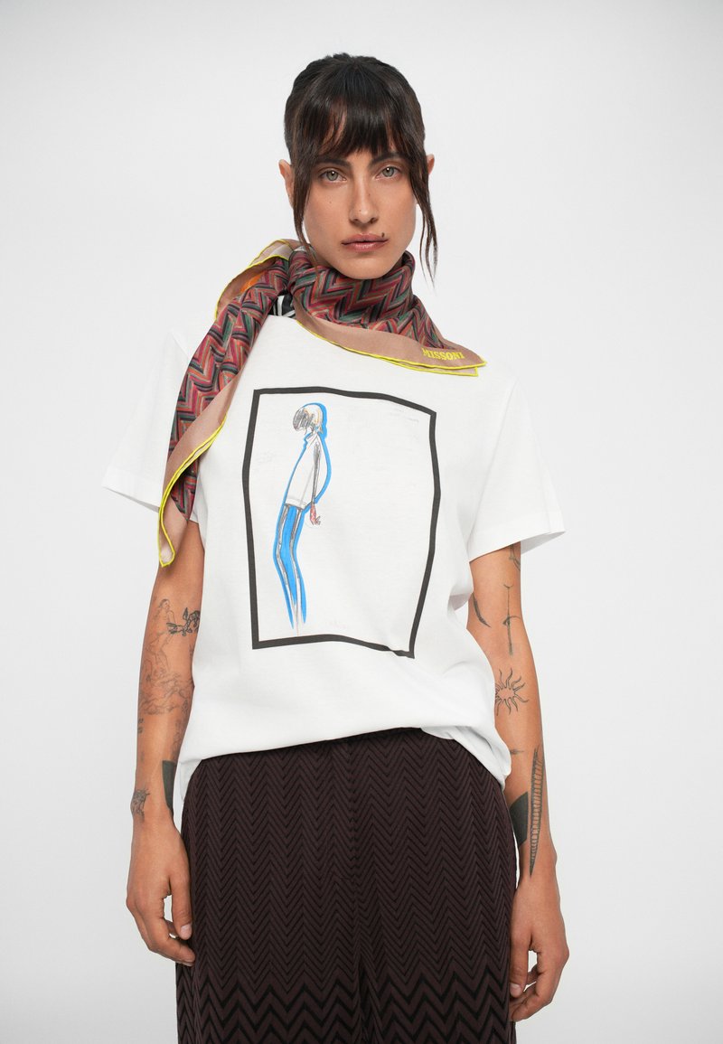 T-shirt bianco in cotone con una grafica di una figura incorniciata di nero, abbinato a una sciarpa a motivi marrone e giallo. Pantaloni a zigzag testurizzati.