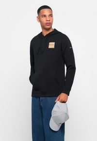 Felpa nera con tasca a marsupio, scollatura rotonda e una toppa logata color nocciola. Abbinata a jeans di denim larghi e porta un cappellino grigio chiaro.