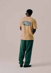 T-shirt en coton beige avec un graphique décoratif vert et blanc au dos, associé à un pantalon de survêtement vert et des chaussures noires.