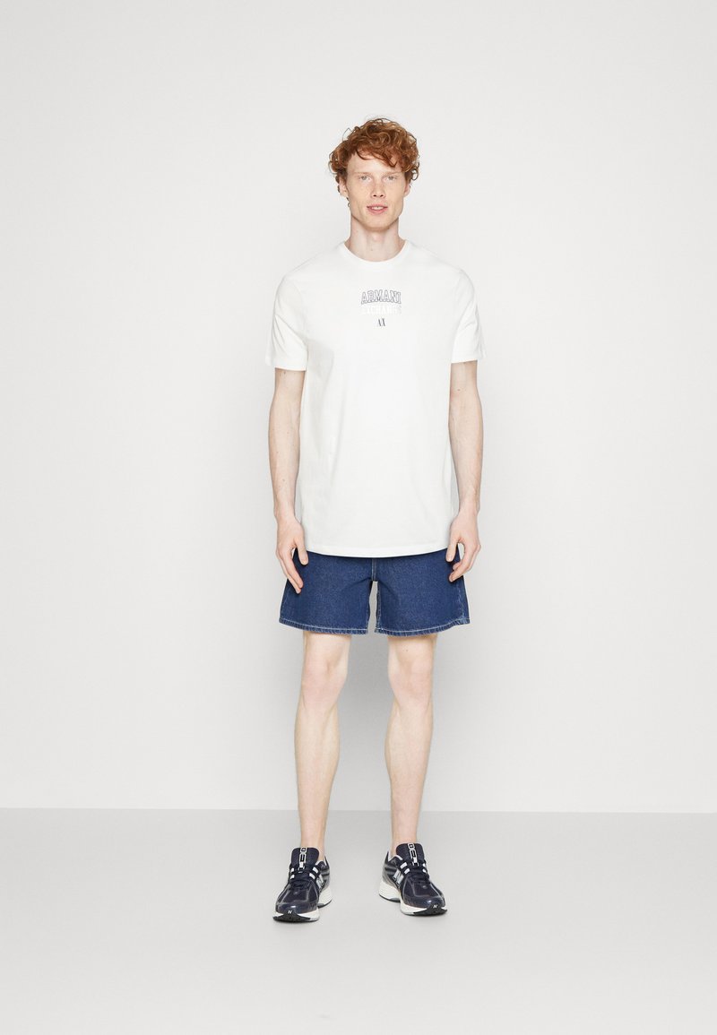 Camiseta de algodón blanca con impresión de logotipo, combinada con pantalones cortos de mezclilla azul oscuro. El modelo lleva zapatillas deportivas en blanco y negro. Conjunto sencillo y casual.