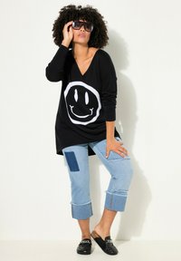 Maglietta nera a maniche lunghe con una stampa di un sorriso bianco, abbinata a jeans cropped azzurri chiari con toppe e orli sfrangiati. Scarpe nere senza lacci.