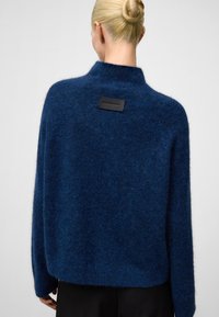 Granatowy sweter o teksturowanej strukturze z wysokim kołnierzem, luźnym kroju i metką z tyłu. Wykonany z miękkiego, puszystego materiału, oferującego swobodny wygląd.
