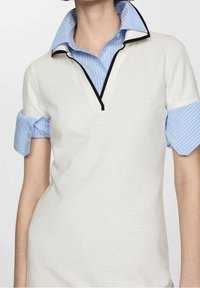 Polo blanc à manches courtes avec un col en V et des bordures noires. Col de chemise à rayures bleu clair dépassant sous les manches du polo.