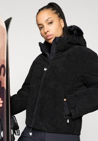Černá kordurová bunda s vysokým límcem a kapucí podšitou kožešinou, se zipovými kapsami a sportovním střihem, vystavená vedle lyží.