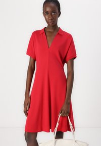 Robe rouge à manches courtes avec un décolleté en V, fabriquée en tissu doux. Silhouette fluide avec une coupe décontractée, associée à un sac à main blanc.