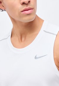 Άνδρας με λευκό αμάνικο αθλητικό μπλουζάκι με γκρι λογότυπο Nike στο στήθος, στραμμένος ελαφρώς προς τα πίσω σε μονόχρωμο φόντο.