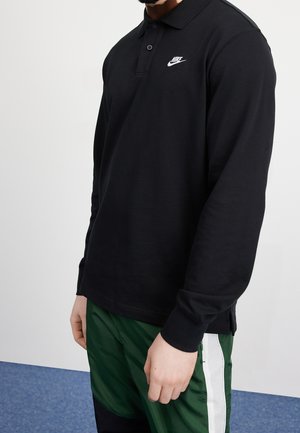 Schwarzes langärmeliges Poloshirt aus Baumwolle mit klassischem Kragen und weißem Nike-Logo auf der linken Brust; kombiniert mit grünen Hosen.